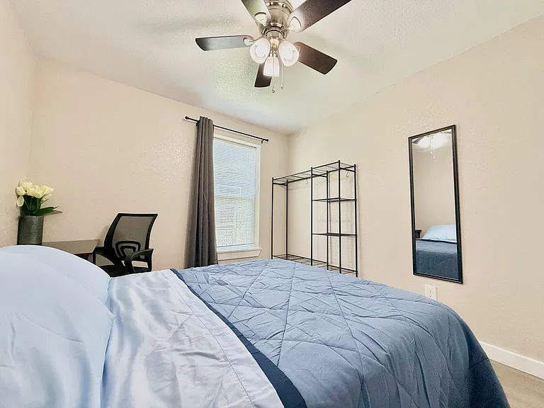 Furnished bedroom with ceiling fan, window with blinds and drapes, open shelving/closet rack, and a work desk area.