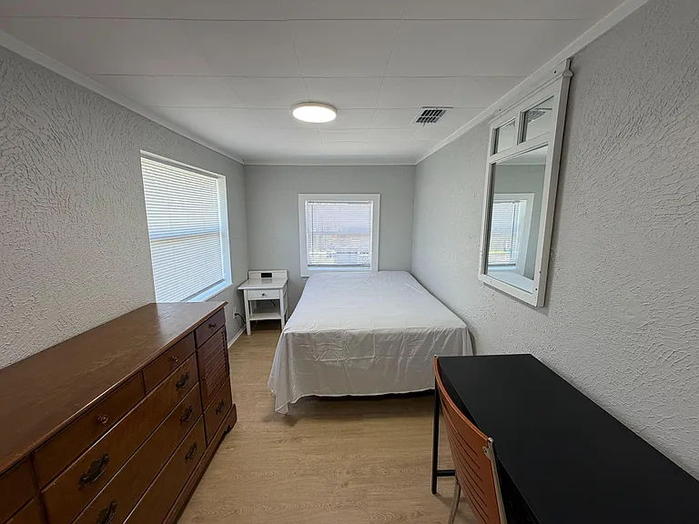 Furnished bedroom with a full-size bed, dresser, and desk, plus two windows with blinds for natural light and privacy.