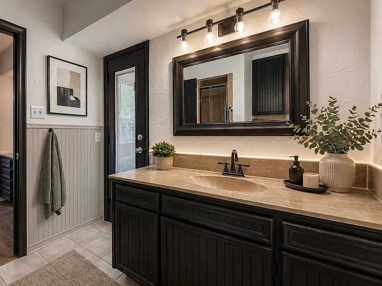 Vanity area with ample counter space, large framed mirror, and storage cabinetry plus a door with window blinds.