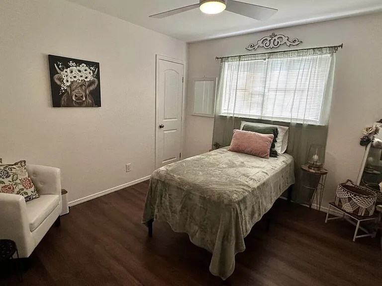 Furnished private bedroom with a single bed, side table, accent chair, and a window with blinds for natural light.