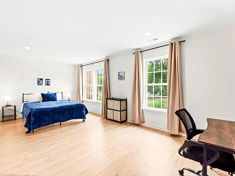 Spacious bedroom with a queen bed, work desk, and two large windows with curtains for natural light and outdoor views.