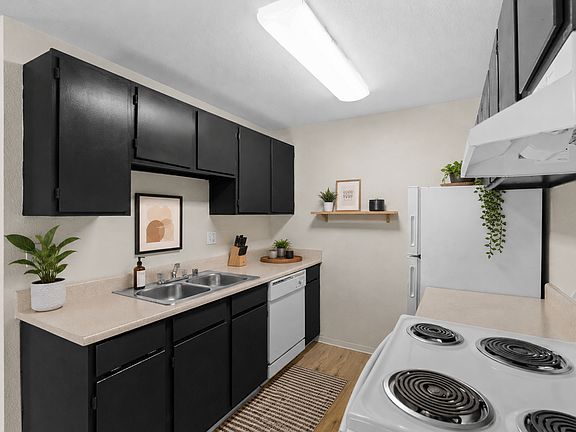 Staged galley-style kitchen with dark cabinetry, light countertops, double-basin sink, dishwasher, electric range, and full-size refrigerator at Garden Square Apartments in Lakewood, WA.