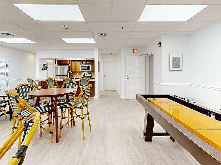 Vibrant community room featuring elegant wicker chairs, a sleek wooden table, and a modern kitchen with stainless steel appliances. Ideal for social gatherings and entertainment.