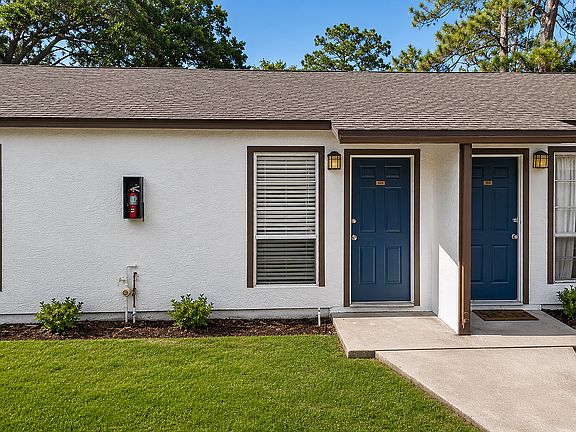 Charming duplex with dual blue doors, manicured lawns, and inviting windows, surrounded by lush greenery.