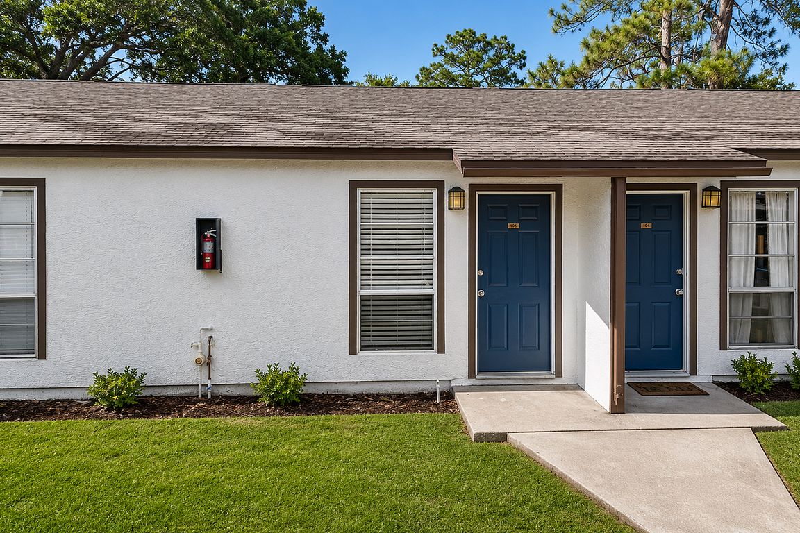 Charming duplex with dual blue doors, manicured lawns, and inviting windows, surrounded by lush greenery.