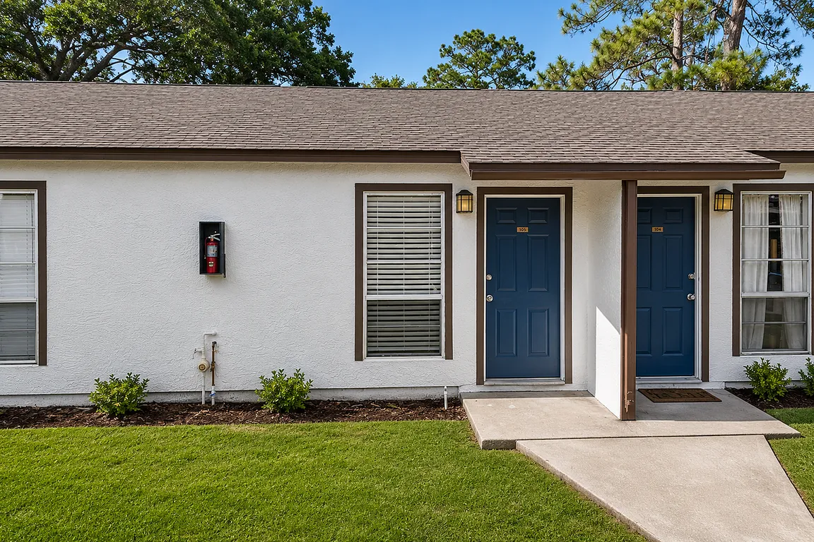 Charming duplex with dual blue doors, manicured lawns, and inviting windows, surrounded by lush greenery.