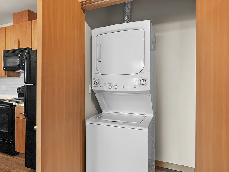 Modern apartment featuring a sleek, in-unit stacked washer and dryer set, efficient kitchen appliances, and warm wooden cabinetry. Modern apartment featuring a sleek, in-unit stacked washer and dryer set, efficient kitchen appliances, and warm wooden cabi