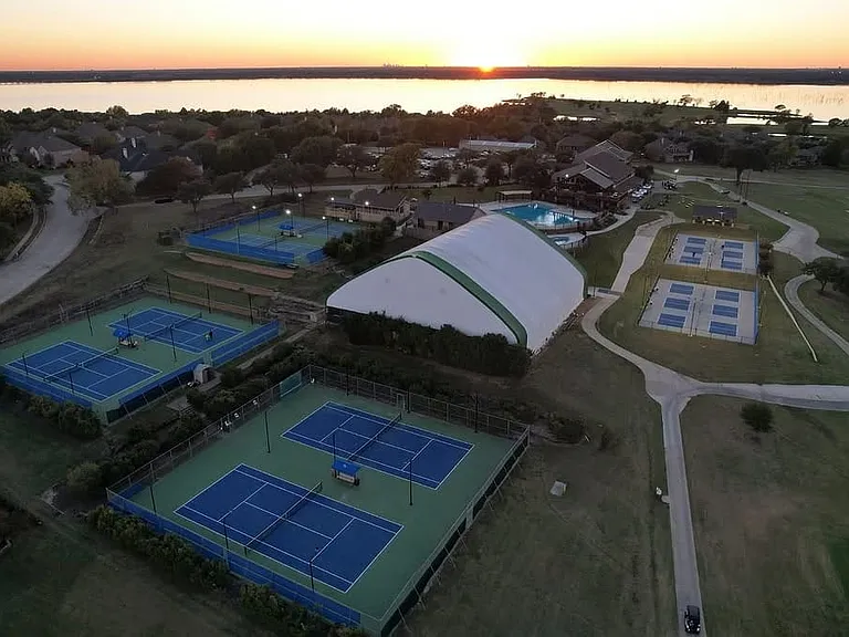 Aerial perspective highlighting multiple outdoor tennis courts, a large indoor court facility, a swimming pool area, and a golf course, all situated alongside a significant body of water