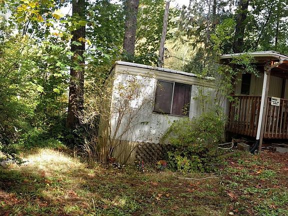 Panoramic photo showing tool shed on the left, the gravel parking area and the front of the trailer with covered porch and addition on the right side. (There is a flaw in the center of the photo that makes it look like there is a separation between the po