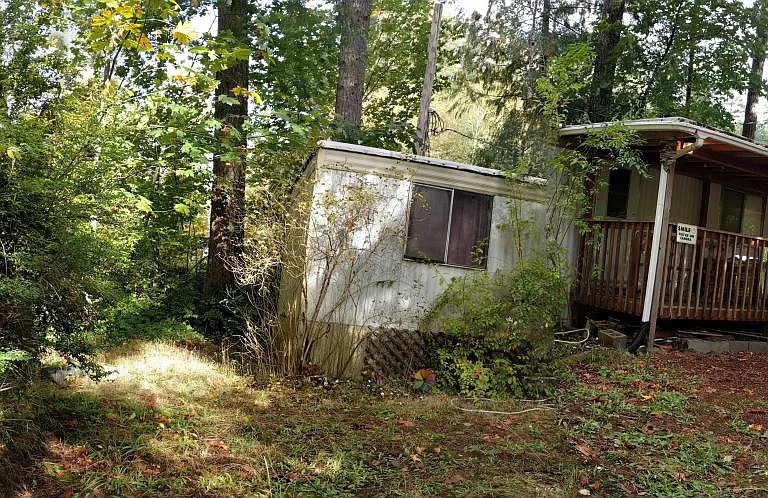 Panoramic photo showing tool shed on the left, the gravel parking area and the front of the trailer with covered porch and addition on the right side. (There is a flaw in the center of the photo that makes it look like there is a separation between the po
