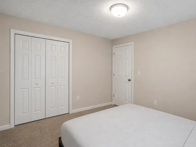 Carpeted bedroom with neutral walls, a wide double-door closet, and space for a full or queen bed and side table.