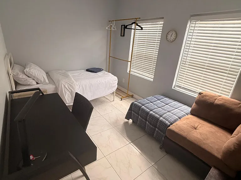 Furnished bedroom with a bed, desk, and lounge seating, plus tile floors and two windows with blinds for light control.