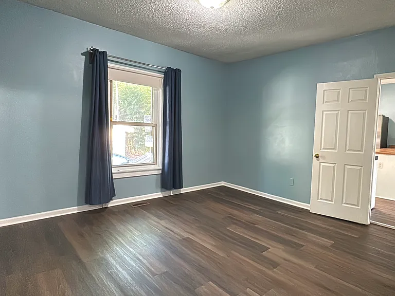 Primary bedroom with calming light blue walls, rich hardwood flooring, and ample natural light from the front window_easily fits a king-size bed and has access to an adjacent private space.