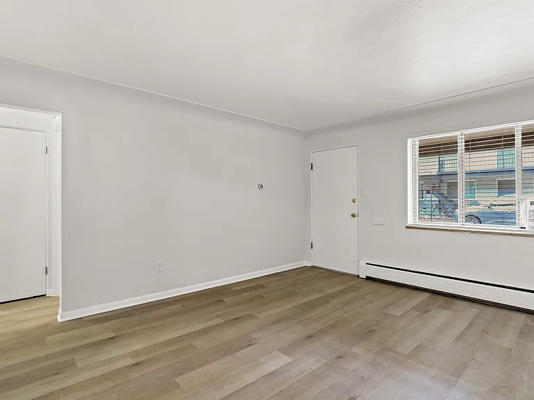 Bright and airy living space with sleek wood flooring, large window for natural light, and access to a cozy room.