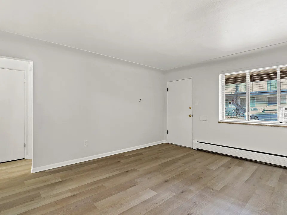 Bright and airy living space with sleek wood flooring, large window for natural light, and access to a cozy room.