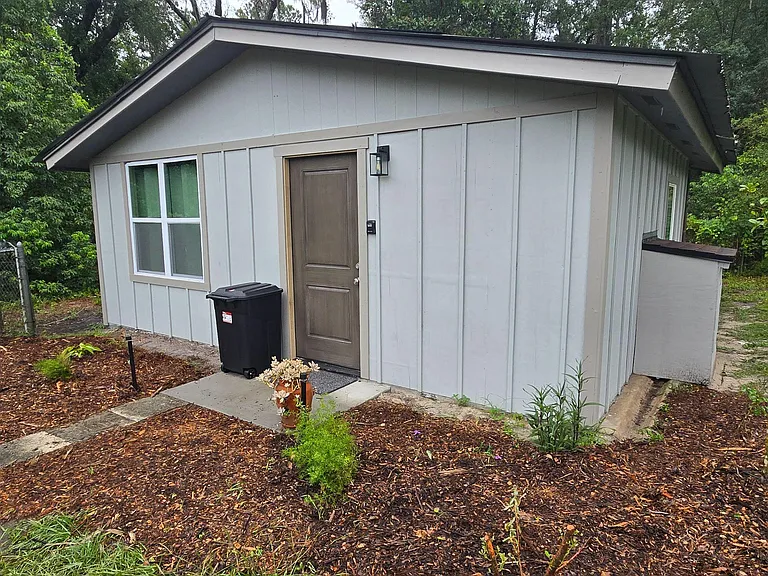 Detached studio apartment with private entry, side windows, and a small landscaped approach set among mature trees.