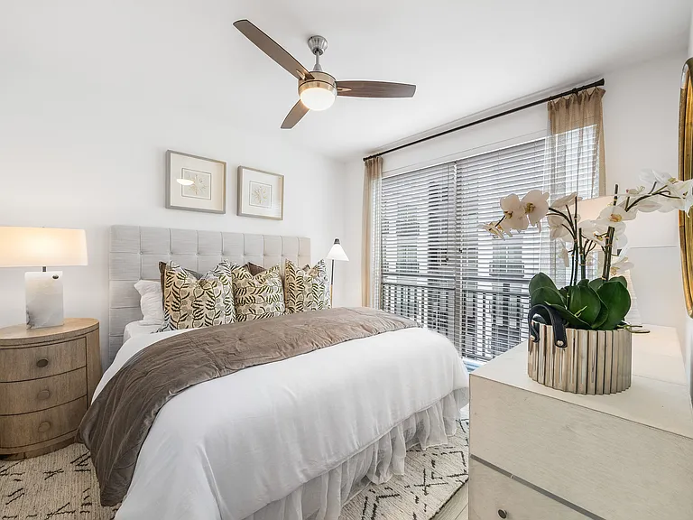 Bright and elegant bedroom featuring a plush bed with patterned pillows, modern ceiling fan, and expansive window with stylish blinds.