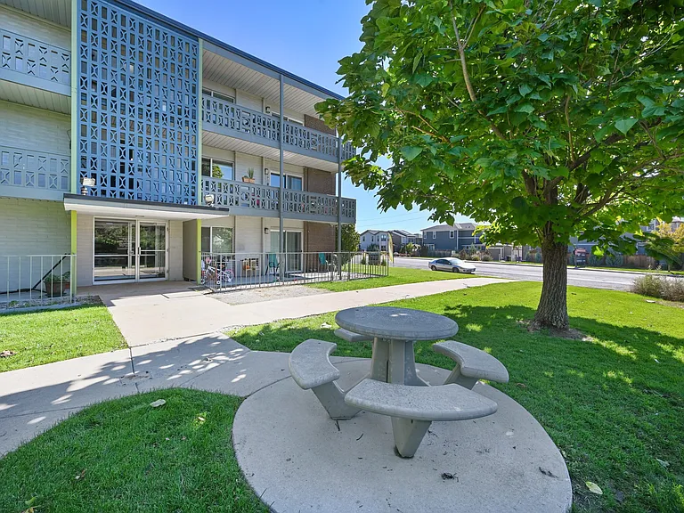 Charming apartment exterior featuring modern architectural design, lush green lawns, and inviting picnic areas underneath shady trees.