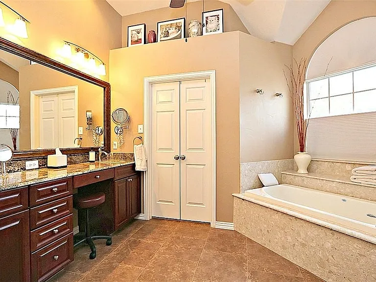 Bathroom featuring a deep soaking tub with a tiled surround, a large arched window, and a dual-sink vanity with a granite countertop and dark wood-finish cabinetry