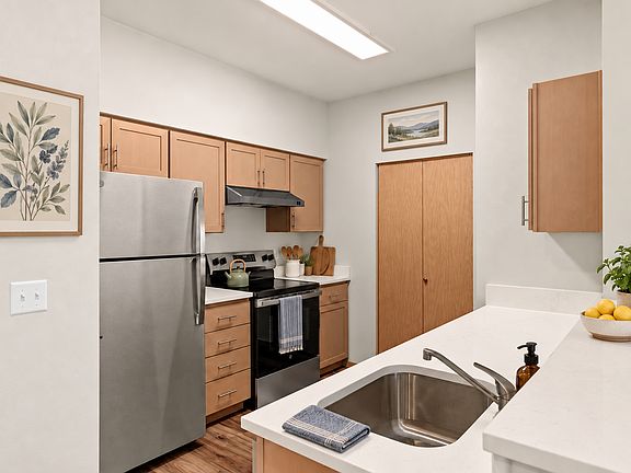 Staged kitchen with stainless steel appliances, light wood cabinets, white countertops, framed artwork, and greenery.