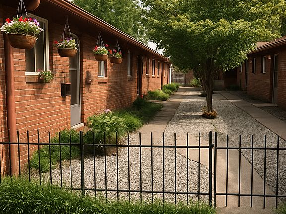 This photo captures a peaceful courtyard between single-story brick apartment buildings. A central sidewalk, black metal gate, hanging flower baskets, gravel landscaping, and a mature tree create a welcoming and well-maintained outdoor space.