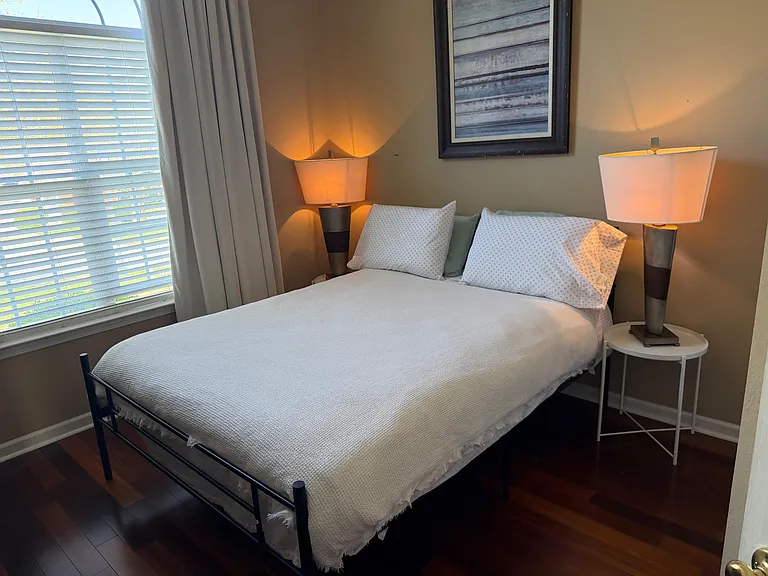 Furnished bedroom with a full-size bed, bedside lighting, and a large window with blinds for adjustable privacy.