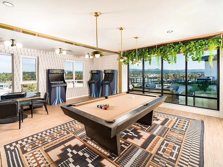 A game lounge at Modera Beaverton with a pool table, arcade games, and large windows that bring in daylight and views beyond the room.