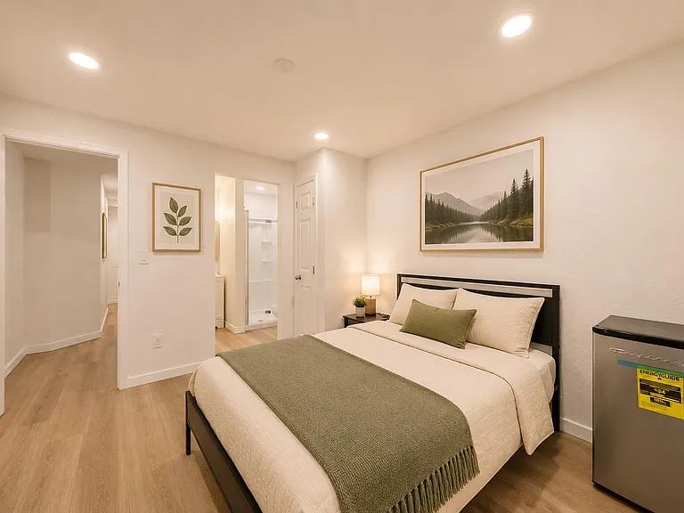 Furnished bedroom with a full bed, nightstand lighting, and a mini fridge, plus easy access to the nearby bathroom.