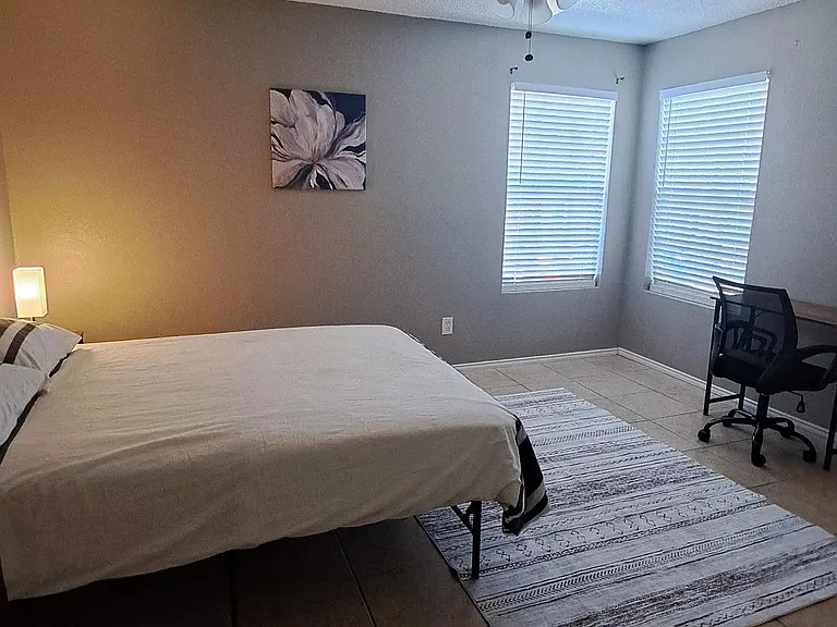 Spacious bedroom with tile flooring, ceiling fan, and two windows with blinds, plus a dedicated desk area for work or study.