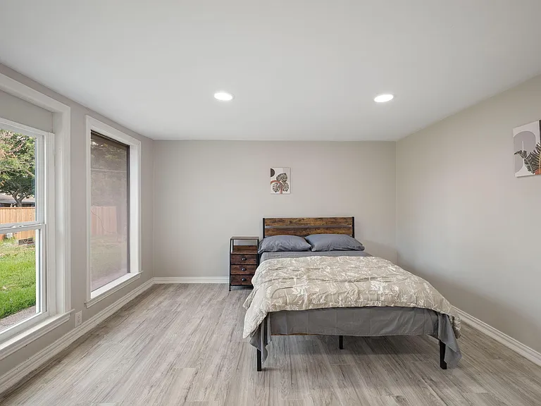 Spacious bedroom with large windows, wood-look flooring, and a comfortable bed setup with a bedside table.
