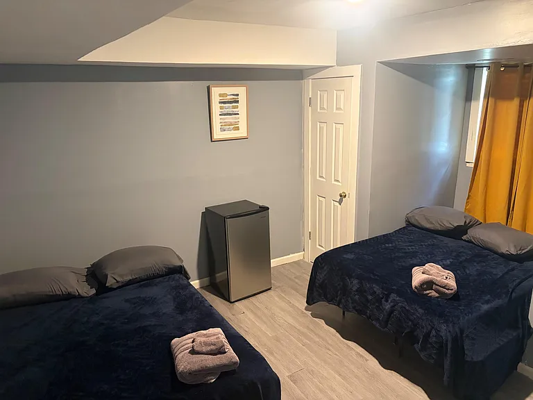 Furnished bedroom with two twin beds, durable flooring, a mini fridge, and a window with curtains for privacy.