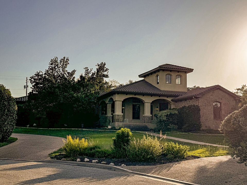 Front elevation at golden hour _ Mediterranean-inspired facade with barrel-tile roof, stone accents, and a wrap-around archway porch that overlooks the lawn and driveways.