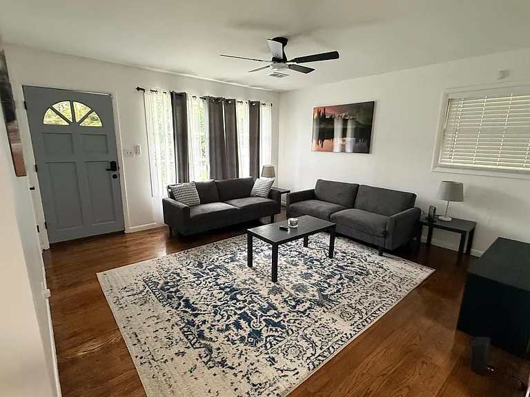 Living room with hardwood floors, two sofas, ceiling fan, and large windows with blinds and curtains for natural light.