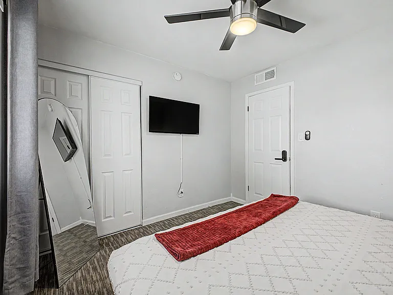 Bedroom features a closet with sliding doors, ceiling fan, and wall-mounted TV, with nearby window blinds for light control.