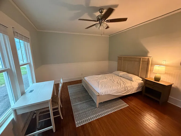 Furnished bedroom with a ceiling fan, hardwood floors, and a desk workspace beside large windows for natural light.