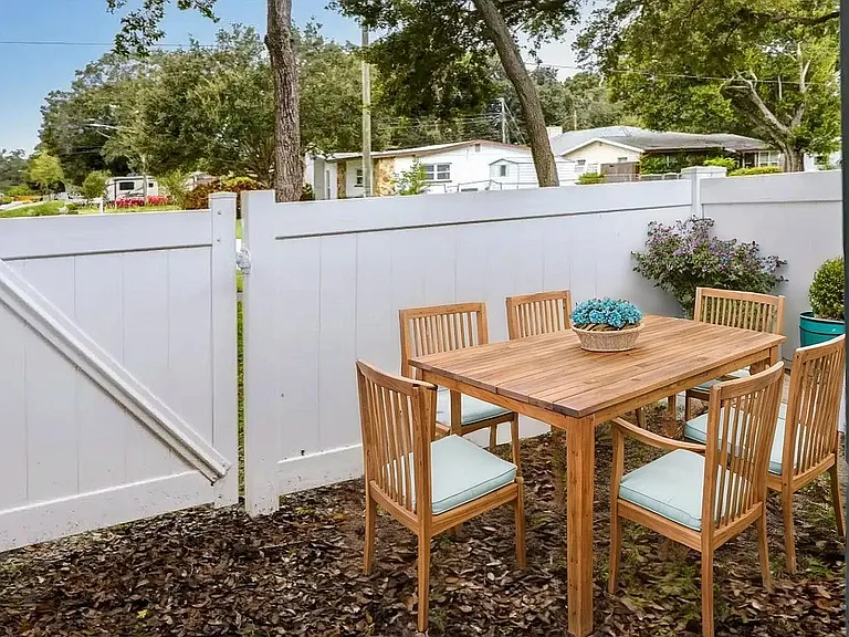 Charming outdoor patio with a wooden dining set for six, surrounded by lush greenery and a cozy white fence, perfect for relaxing and entertaining.