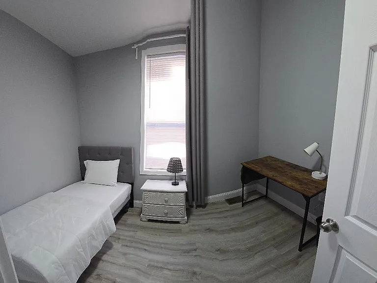 Private bedroom with twin bed, bedside storage, and a work desk; window with curtains provides natural light.