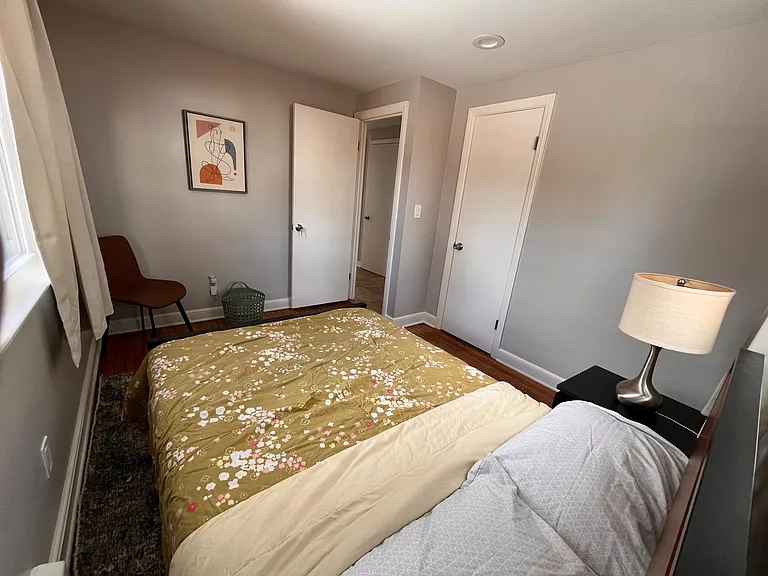 Furnished bedroom with hardwood floors, window with curtains, bedside table and lamp, plus a closet for storage.