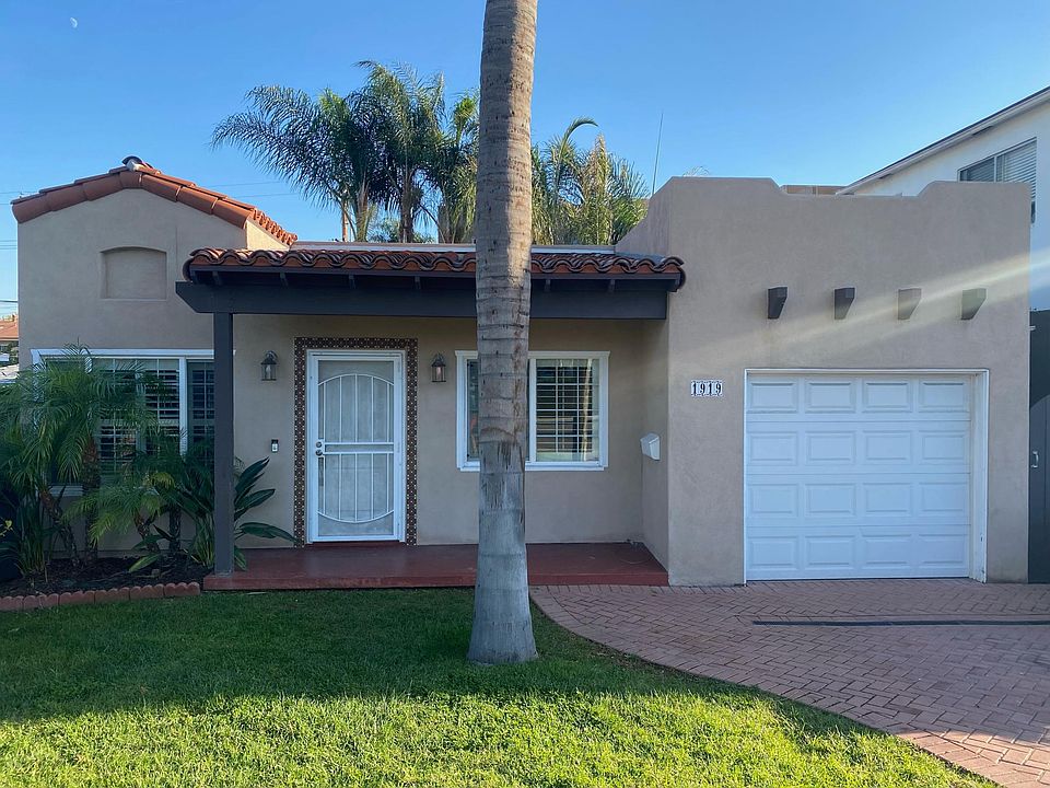 Charming Spanish-style house with mature palm trees, grassy lawn, and a brick pavestone driveway.
