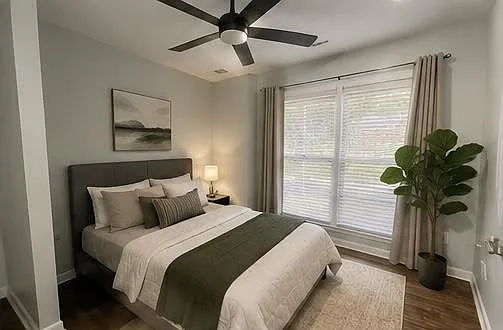 Furnished bedroom with a ceiling fan, large window with curtains, bedside lighting, and space for a full/queen bed.
