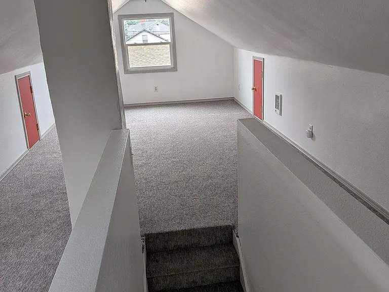 Carpeted upper-level landing with sloped ceilings, overhead lighting, and a window bringing in natural light.