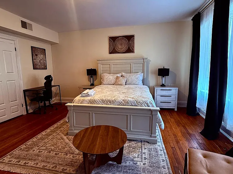 Furnished bedroom with a full bed, nightstands, and a dedicated desk area, plus hardwood floors and large windows.