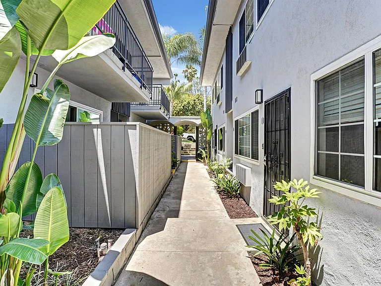 A concrete pathway leads between two rows of plants and a white building with black screen doors.