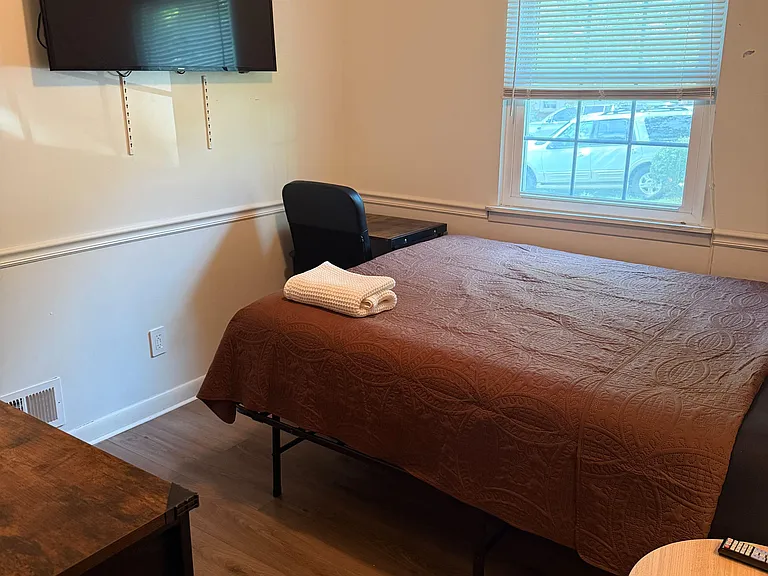 Furnished bedroom with a comfortable bed, wall-mounted TV, and a window with blinds for natural light and privacy.