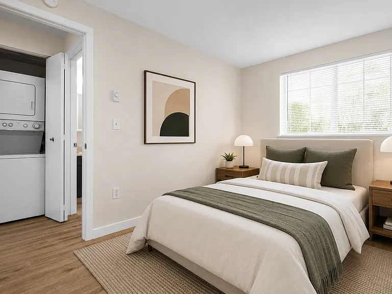 Staged bedroom with wood-style plank flooring, large window with blinds, natural light, and direct access to in-unit stacked washer and dryer at Garden Square Apartments in Lakewood, WA. Staged bedroom with wood-style plank flooring, large window with bli