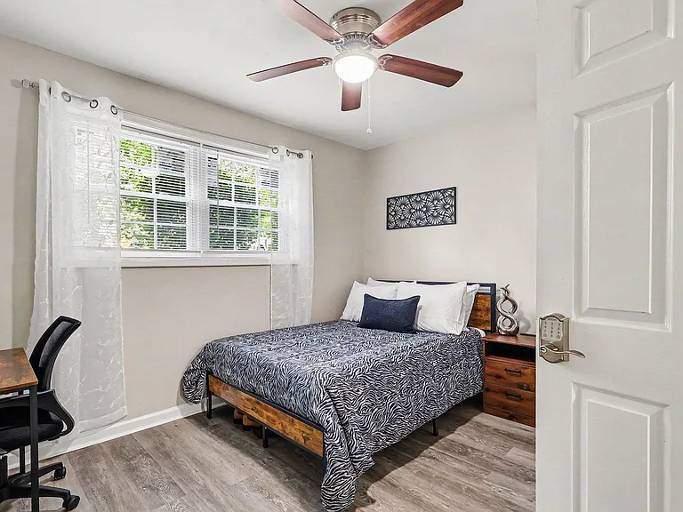 Furnished bedroom with a full-size bed, nightstand, and desk area, plus a large window and ceiling fan for comfort.