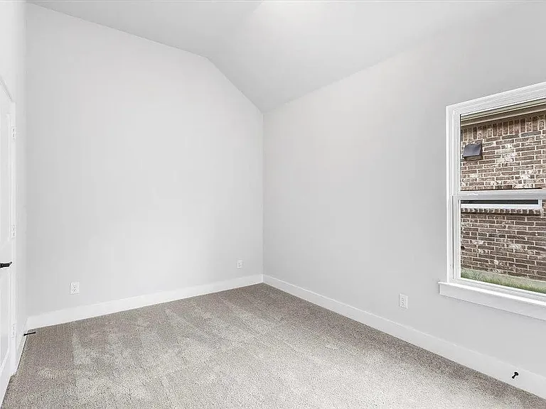 Vaulted ceiling bedroom with light gray carpeting, white baseboards, and a single window offering exterior brick wall visuals