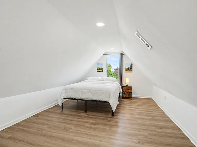 Loft-style bedroom with vaulted ceilings, wood-look flooring, and a large window with curtains for natural light.