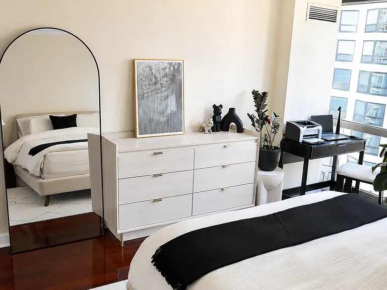 Another angle of the bedroom highlighting crisp walls and dark hardwood floors. The layout offers plenty of room for a full dresser, a large floor mirror, and your office desk setup right by the window without feeling cluttered.