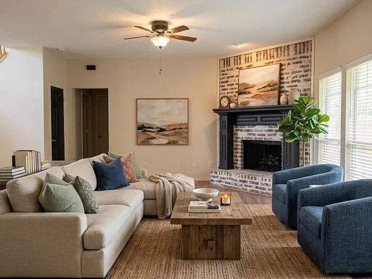 Virtual staging: Living space featuring a whitewashed brick fireplace with a dark wood mantel, wood-finish flooring, two large windows with blinds, a ceiling fan, and a staircase with wood railings